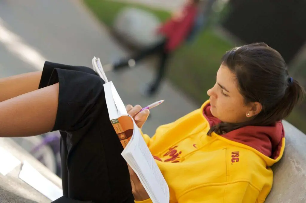 A student in a USC hoodie writes in a notebook outdoors. In this article, learn how to pick a college essay topic.