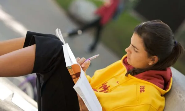 A student in a USC hoodie writes in a notebook outdoors. In this article, learn how to pick a college essay topic.