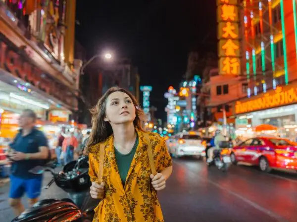 A young woman wearing a backpack walks through the city streets of Bangkok, Thailand. Learn about the pros and cons of taking a gap year.