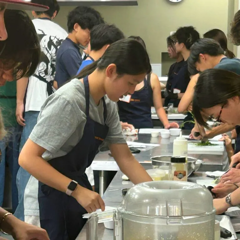 A view down a long, stainless steel table at which students prepare pasta sauce ingredients, including pulsing garlic in a food processor.