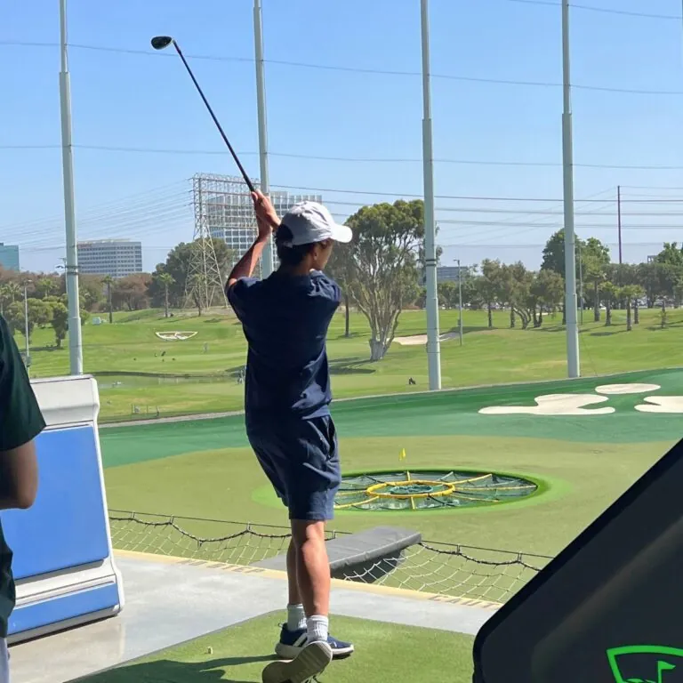 A student swings a golf club at a driving range while another watches.