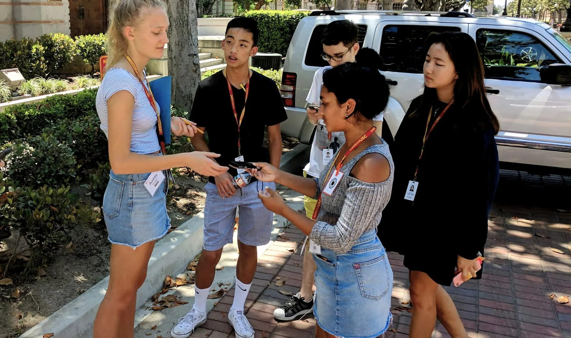 Students stand outdoors discussing notes and using their phones during a group activity.
