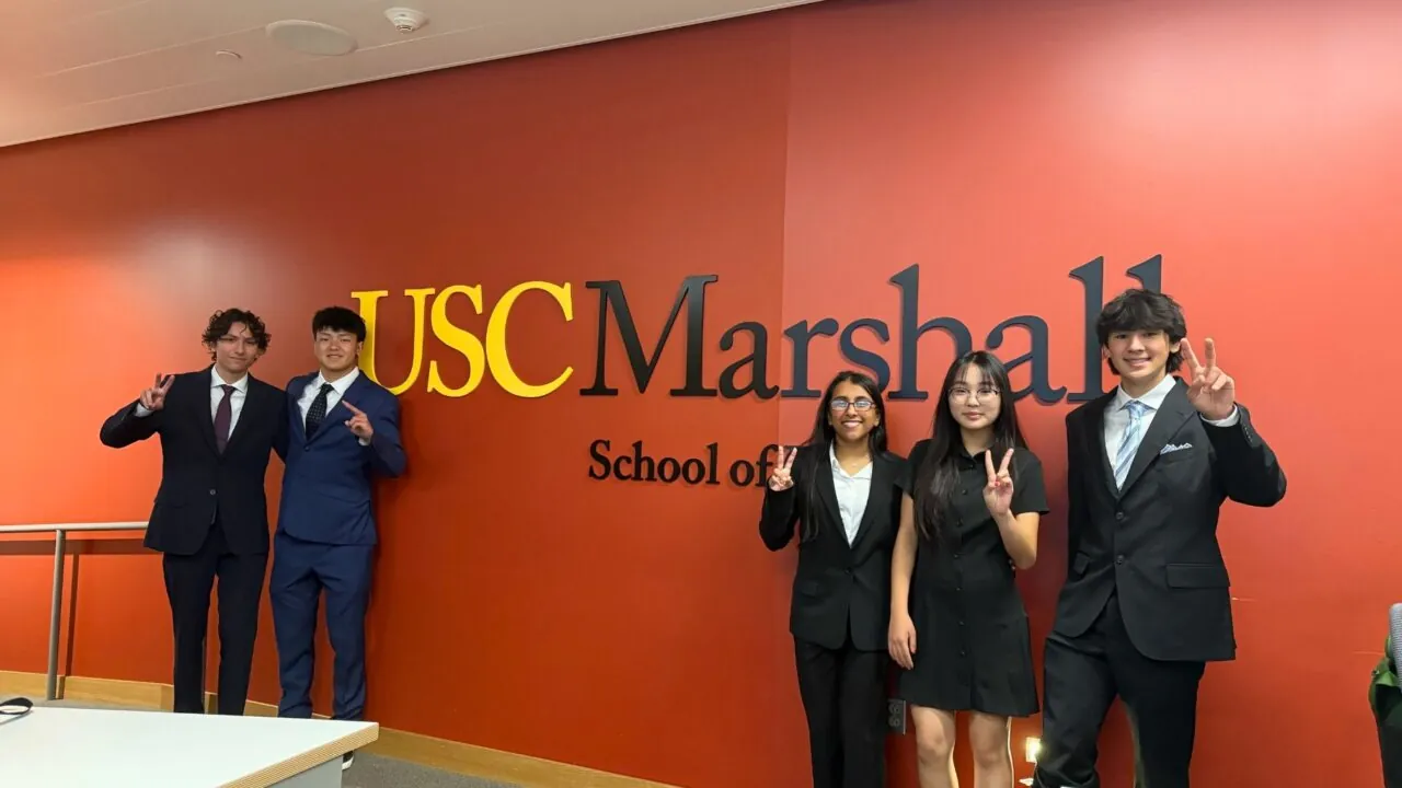 Five students in professional attire stand in front of a red wall with the USC Marshall School of Business logo, holding up the "Fight On" hand sign.
