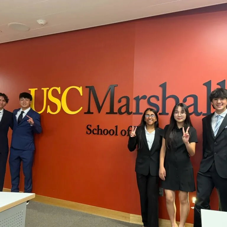 Five students in professional attire stand in front of a red wall with the USC Marshall School of Business logo, holding up the "Fight On" hand sign.