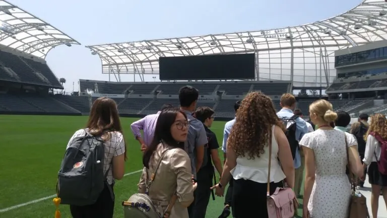 Students walk across the field at an open-air stadium under a white canopy structure, looking around during a tour.