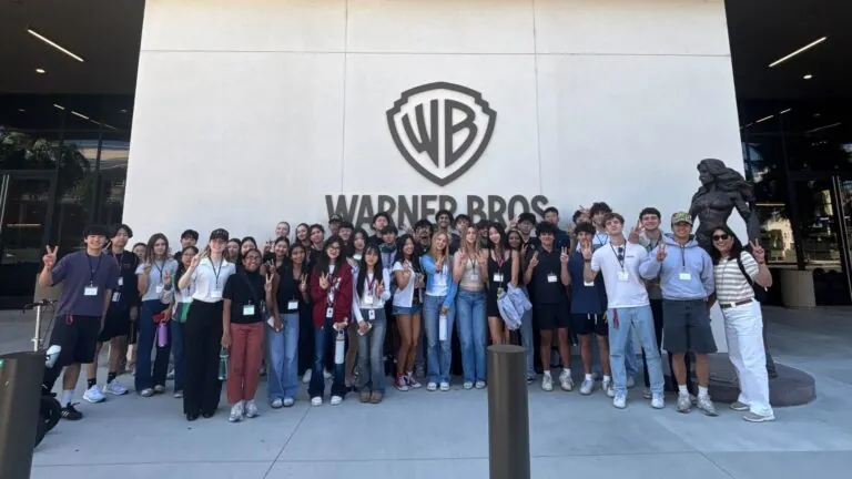A large group of students poses together outside the Warner Bros. Studios building under the “WB” logo.