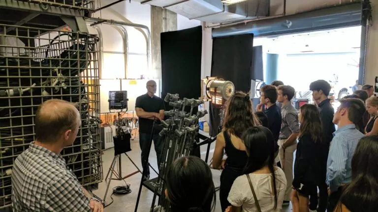 A group of students listens attentively to an instructor in a lighting and production studio filled with professional film equipment.