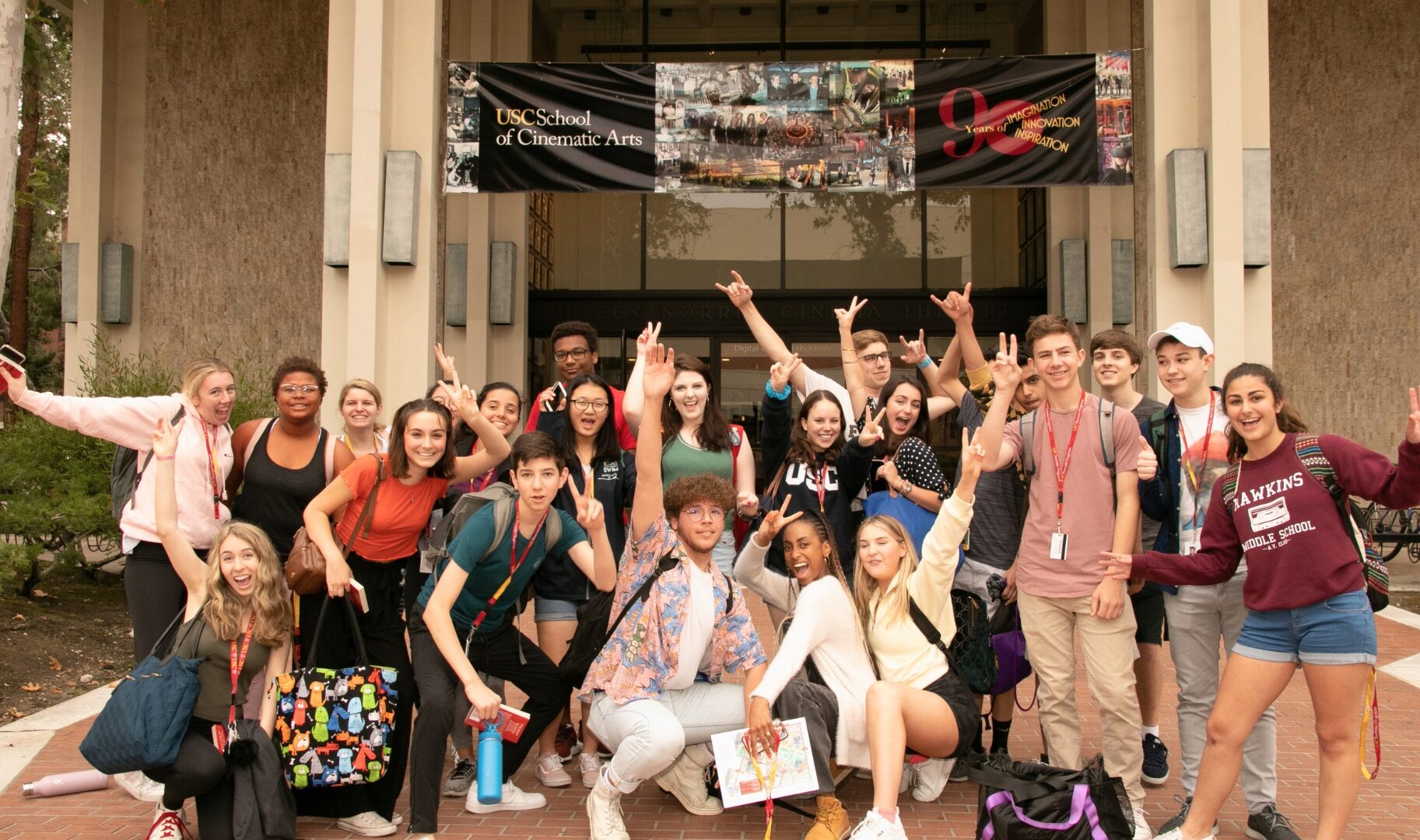 Comedy performance students pose under banner for the USC School of Cinematic Arts on a campus building.