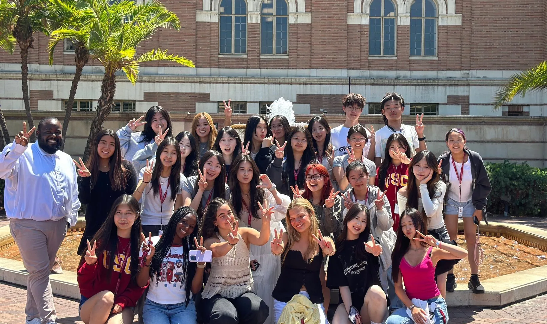 A large group of students pose outside a USC building in front of a fountain, smiling and making peace signs.