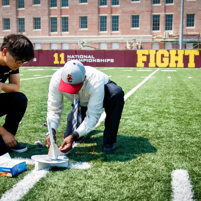 [Photo] Two people kneel on a football field, working together to assemble or adjust a small model rocket on a launch pad near a painted yard line. Stadium seating, a scoreboard and a large "Fight On!" sign are visible in the background under a sunny sky.