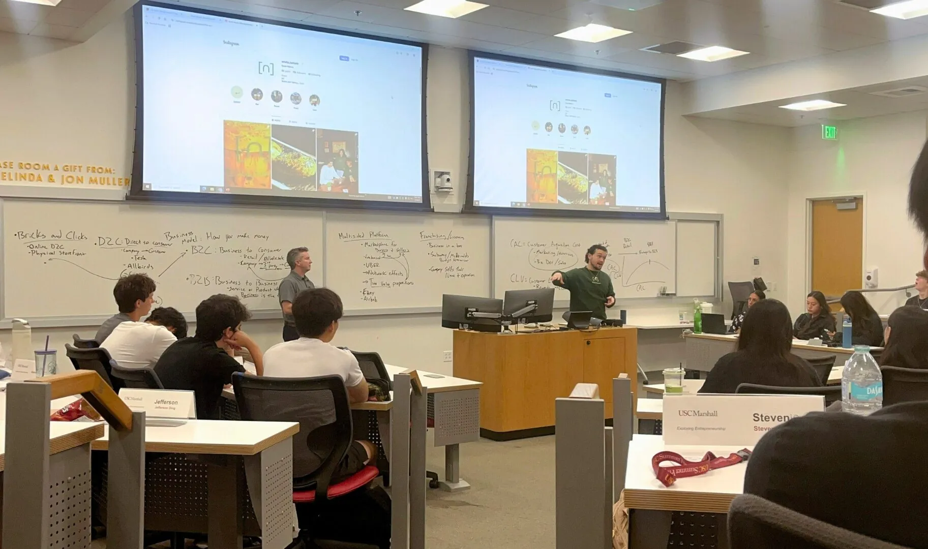 A classroom of students listens attentively as two instructors present at the front of the room, with slides and whiteboard notes about business models and marketing strategies displayed behind them.