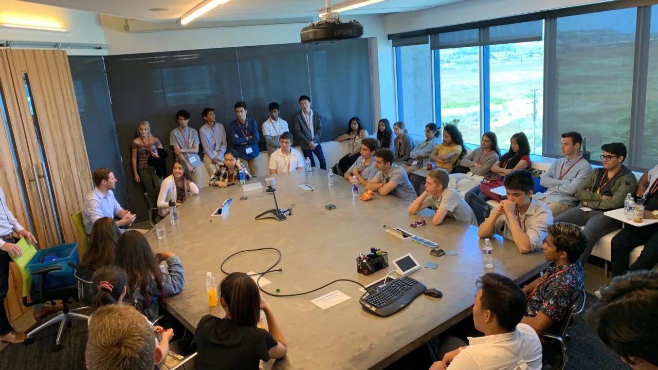 A large group of students sits around a conference table listening to a professional speaker during a company visit, with panoramic windows providing a view of the cityscape outside.