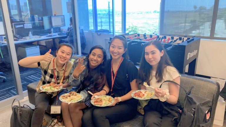 Four students sit together indoors, smiling and holding plates of food during a casual lunch, with computers and a foosball table in the background.