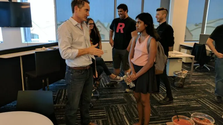 A professional speaks one-on-one with a student holding a water bottle, while other participants in the background share a laugh during an informal networking event.