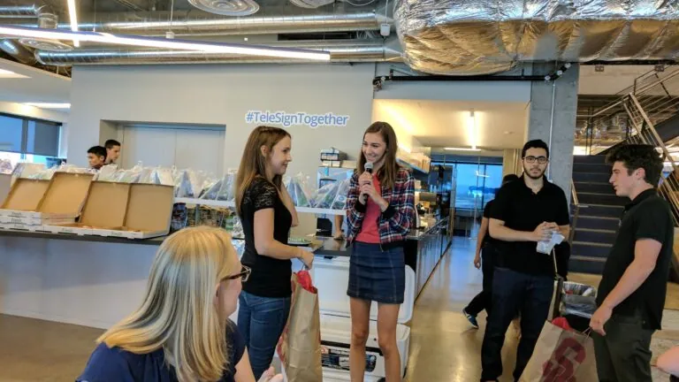 A student with a microphone speaks to another person during a casual office gathering, while others look on and share a laugh under a sign that reads “#TeleSignTogether.”