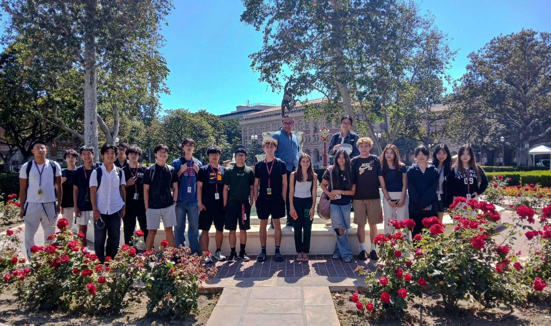A group of students posing for a photo in front of a fountain surrounded by red flowers on campus.