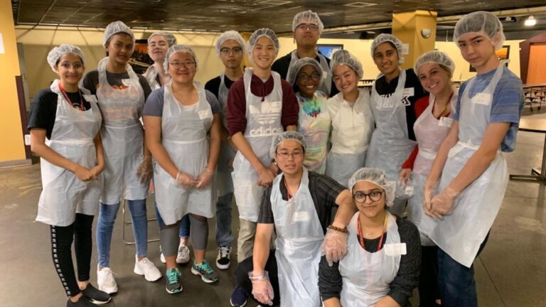 Students wearing hairnets and aprons pose together in a cafeteria setting during a service project.