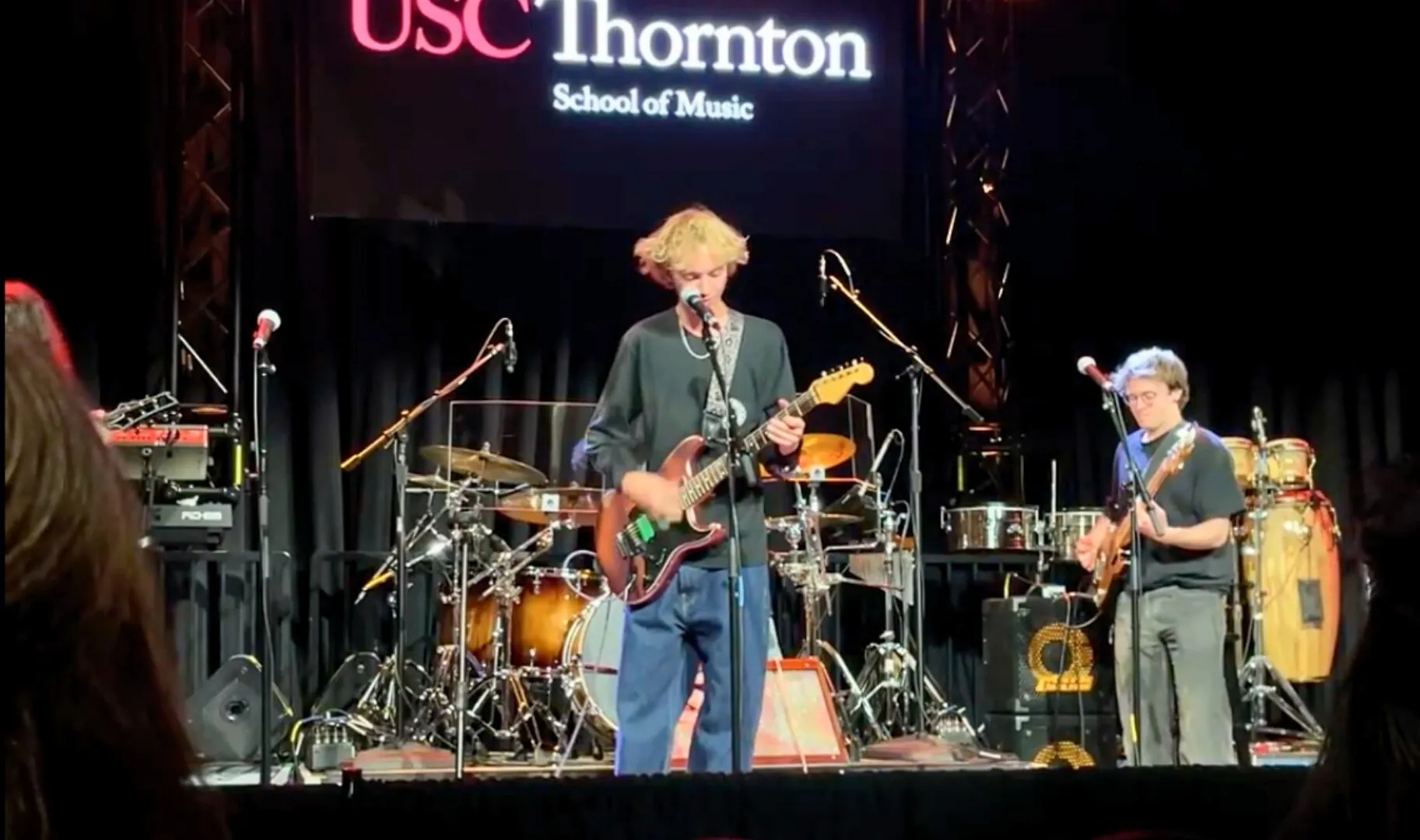 A student performs onstage with an electric guitar during a live concert at USC Thornton School of Music, accompanied by other musicians on drums and bass under stage lights.
