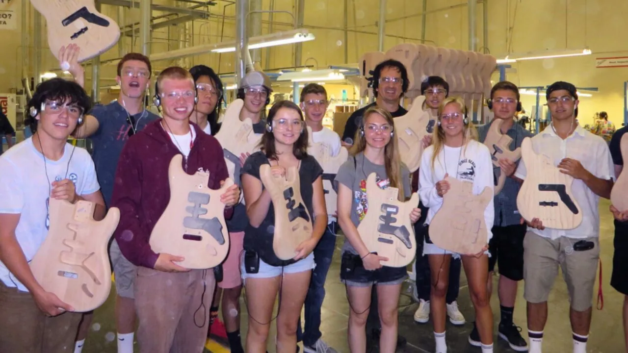 A group of students wearing safety goggles holds unfinished wooden guitar bodies inside a workshop, smiling after a hands-on instrument-building session.