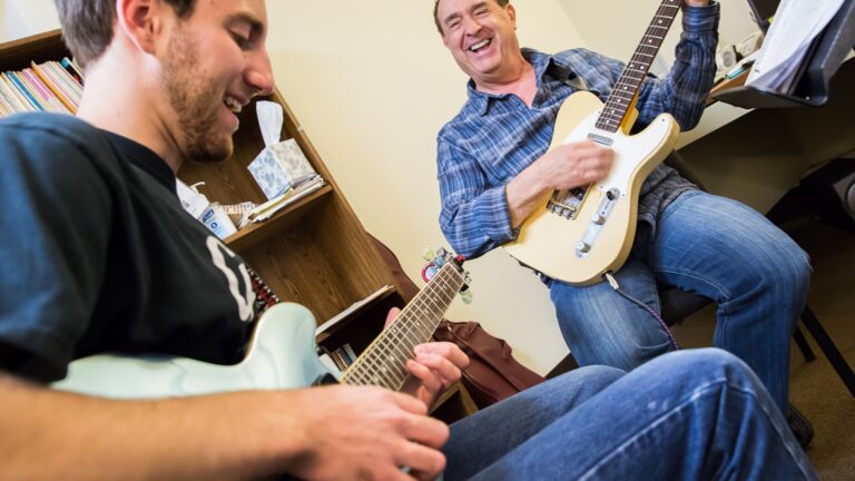 Two people seated in a small music room play electric guitars together, smiling and enjoying the moment during a lesson.