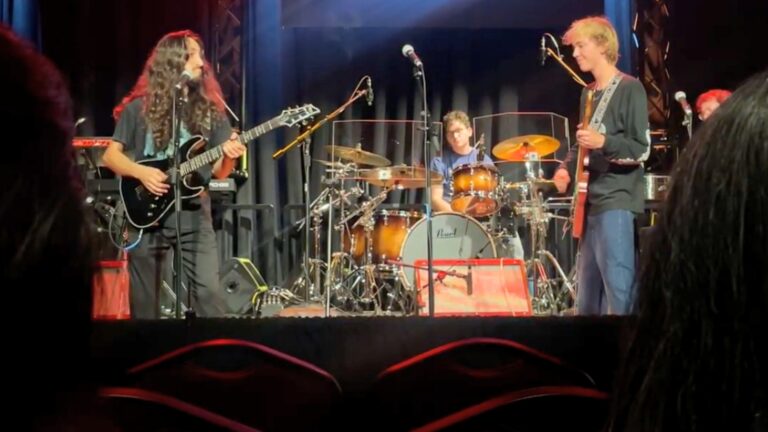 Two guitarists perform on stage with a drummer behind them during a concert at the USC Thornton School of Music, illuminated by stage lights.