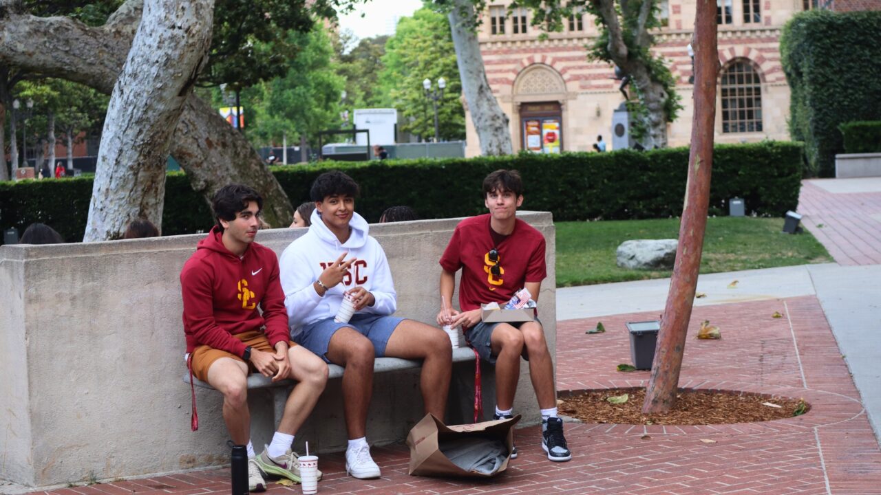 Three students sitting outdoors on a bench, wearing USC sweatshirts and chatting.