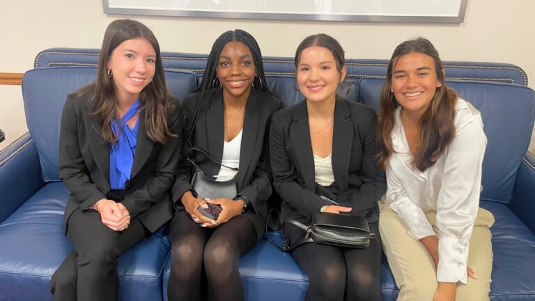 Four students dressed in suits sit together on a blue leather couch, smiling at the camera in a professional setting.