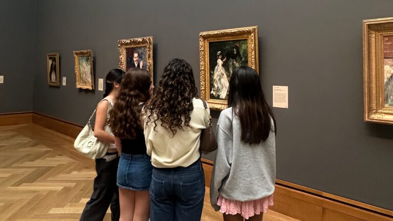 Three girls with their backs to the camera look at a gold-framed painting hanging on a dark wall at The Getty.