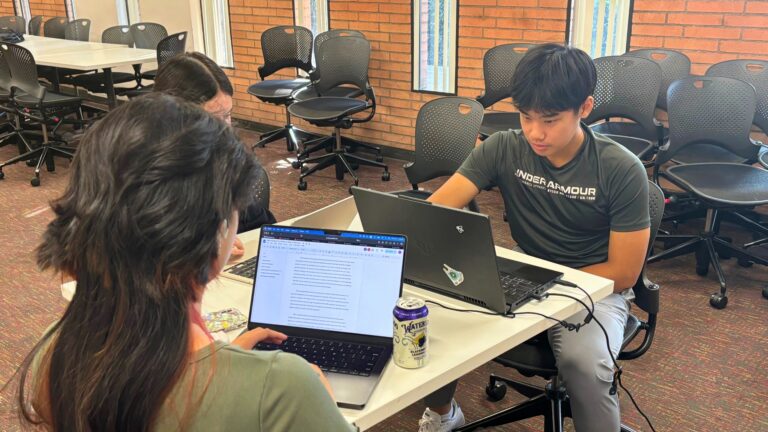 Two students study on their laptops at a table in a classroom with brick walls.