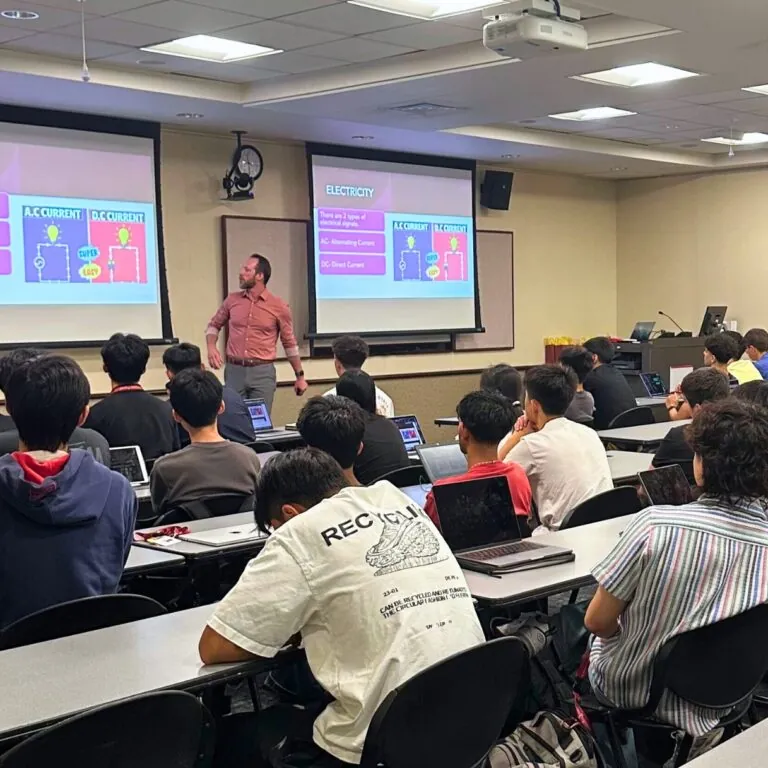Students in a classroom listen to lecture from a professor, who is giving a slideshow presentation.