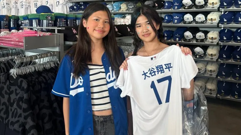 Two girls stand in the gift shop of the Dodger Stadium with rows of baseball caps in the background. One holds up a jersey with the number 17.