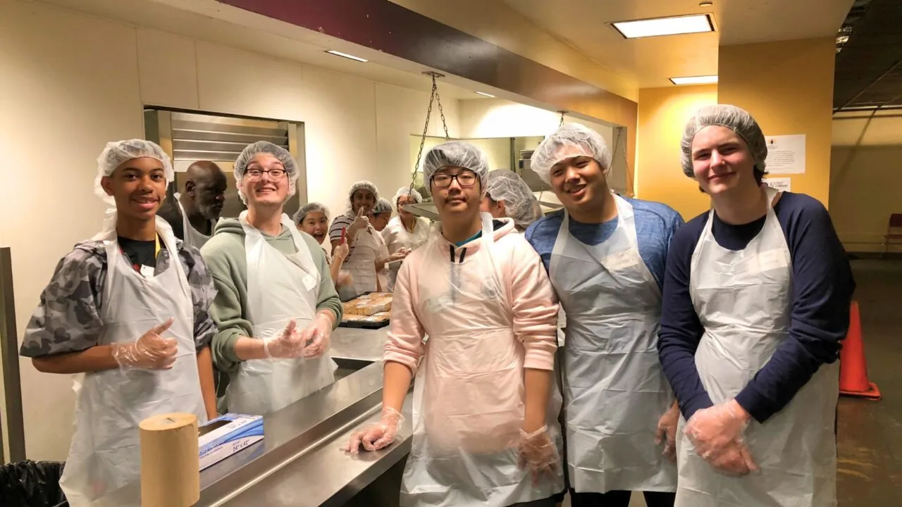 A group of students wearing aprons, gloves and hairnets smiles behind a serving counter while preparing food in a community kitchen.