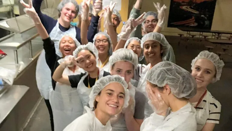 A cheerful group of students wearing aprons and hairnets poses in a kitchen, smiling and raising their hands while volunteering together.