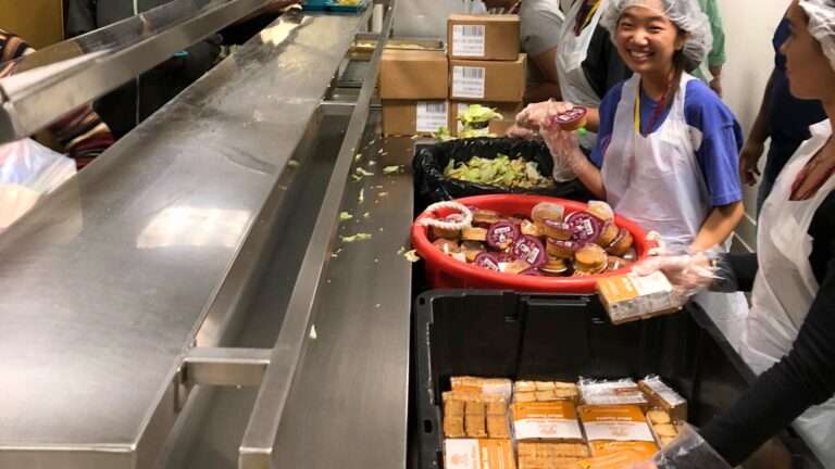 Students wearing aprons and gloves serve packaged meals and salad portions across a cafeteria counter during a volunteer service project.