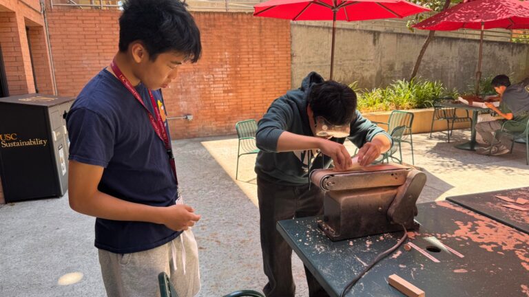 Two students work outdoors sanding a project on a table under red umbrellas.