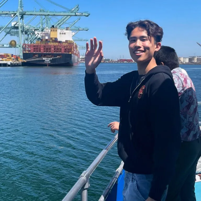 A student waves from a boat near a shipping port with cranes and containers in the background.