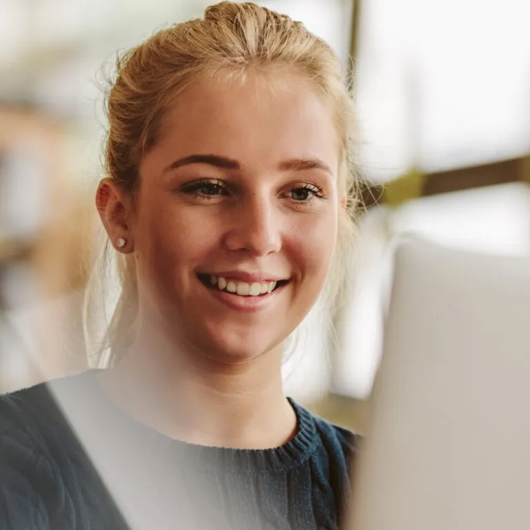 A smiling student looks at a laptop screen while studying in a bright room.