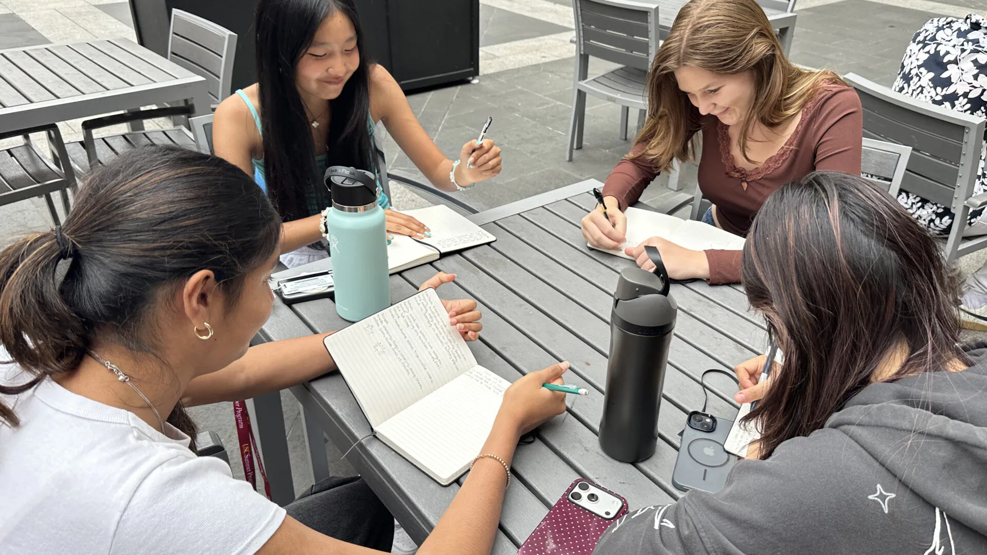 [Photo] Four young women sit around a gray outdoor table, smiling and writing in open notebooks. Water bottles and phones rest on the table amid gray patio furniture.