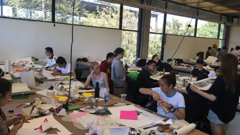 Students work on mixed media projects at large tables in a windowed classroom.