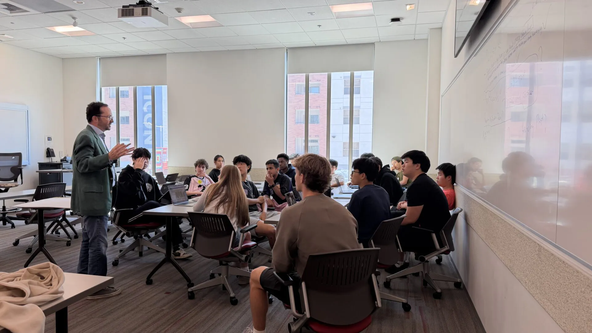 [Photo] An instructor speaks to students seated in small groups around classroom tables while gesturing during a discussion. Large windows bring in daylight as students listen, participate and work on laptops.