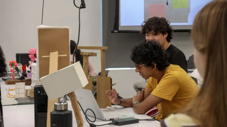 [Photo] Students work at a classroom table with laptops and cardboard prototypes while one student looks at a laptop screen and another smiles from behind. Small handmade models and presentation materials are arranged across the table, reflecting an ongoing design project.