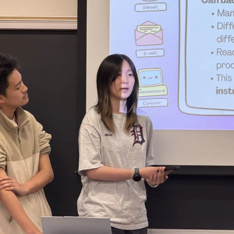 [Photo] Two students stand at the front of a classroom presenting while one speaks and the other listens beside a projected slide. The slide summarizes a study about how background instrumental music may influence reading comprehension and includes simple illustrations of internet, email and computer icons.
