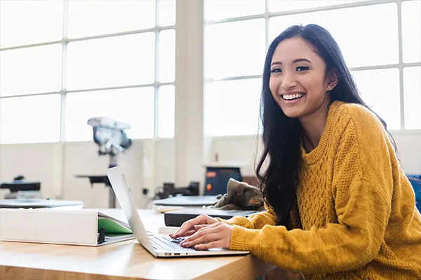 Smiling high school student in a yellow sweater typing on a laptop in a bright classroom, representing USC Pre-College Online Exploration Courses that let students discover academic interests and learn flexibly.