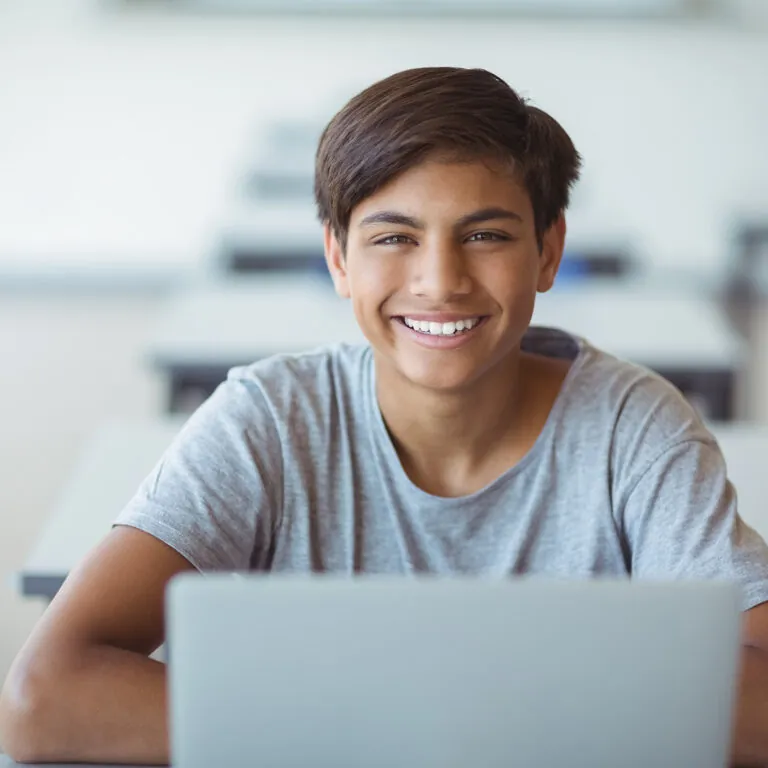 Smiling high school student sitting in a classroom with a laptop, representing USC Pre-College online course “Analytics: The Power of Data for Businesses,” where students learn at their own pace and earn a Certificate of Completion.