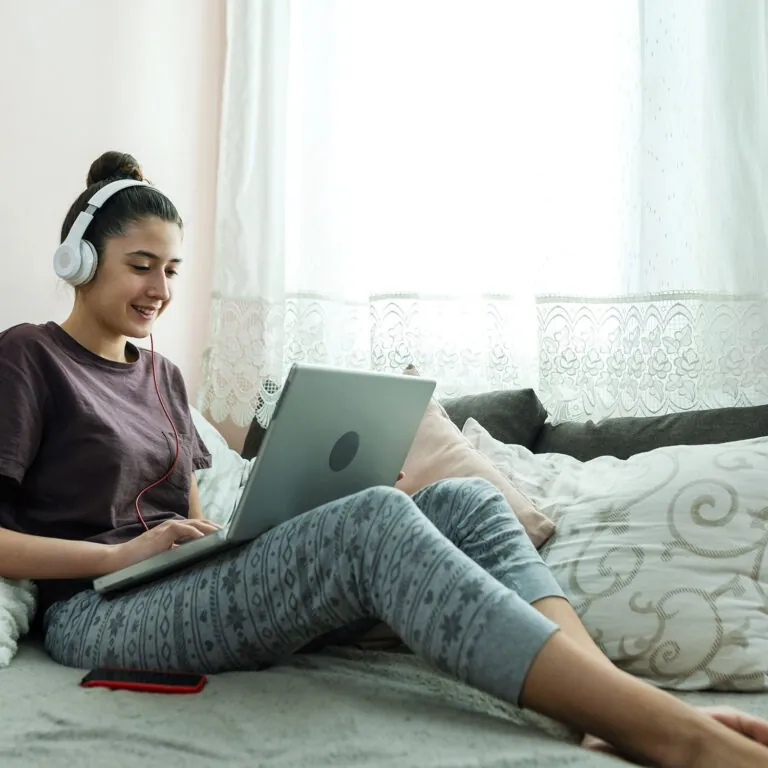 Smiling student wearing headphones and studying on a laptop while sitting on a bed, representing USC Pre-College online course “Environmental Studies: Sustainability and Your World,” where students learn at their own pace and earn a Certificate of Completion.