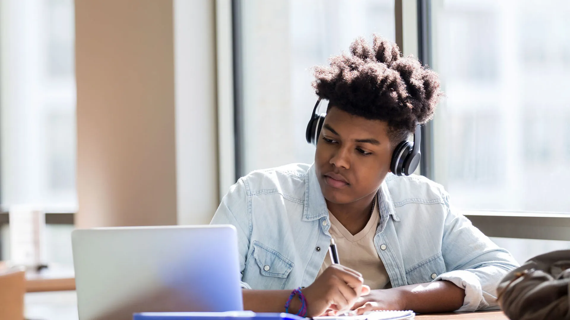 Focused student wearing headphones and taking notes while studying on a laptop near a window, representing USC Pre-College online course “Screenwriting: From Ideation to Writing Your First Script,” where students learn at their own pace and earn a Certificate of Completion.