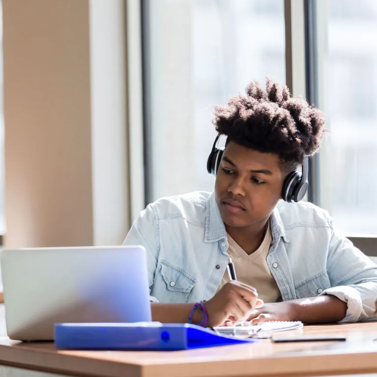 Focused student wearing headphones and taking notes while studying on a laptop near a window, representing USC Pre-College online course “Screenwriting: From Ideation to Writing Your First Script,” where students learn at their own pace and earn a Certificate of Completion.
