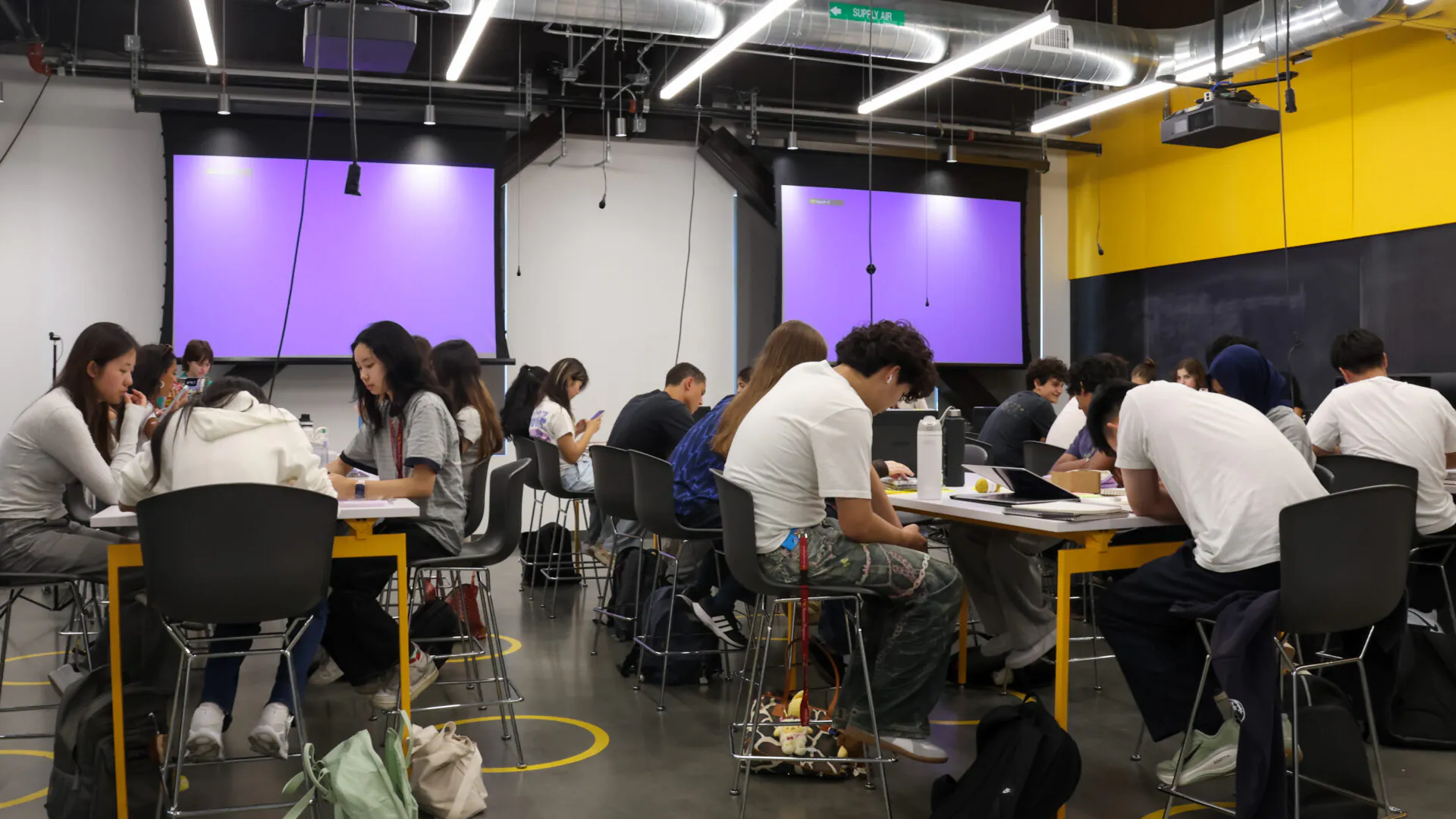 [Photo] Students sit in small groups around high tables, collaborating on classwork in a modern classroom with exposed ceilings, bright overhead lighting and two large purple projection screens at the front. Backpacks, notebooks, laptops and water bottles are placed around the room as students focus on discussion and individual work.